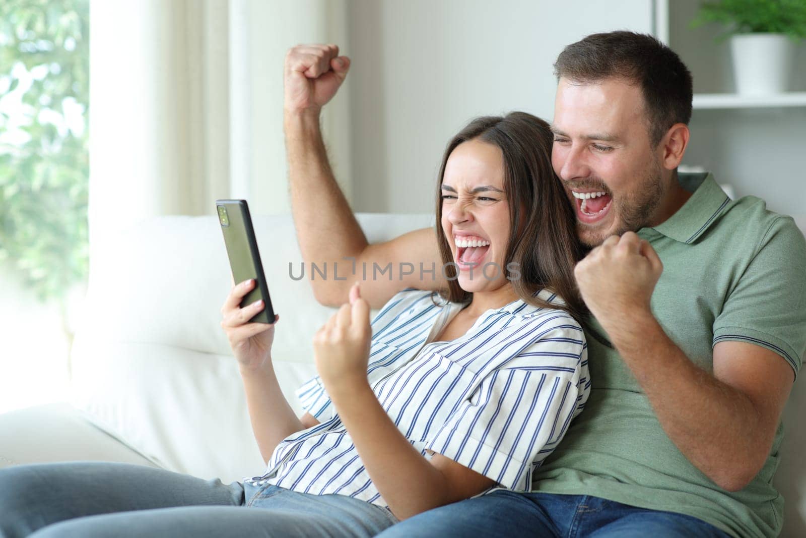 Excited couple checking mobile phone sitting in a sofa at home