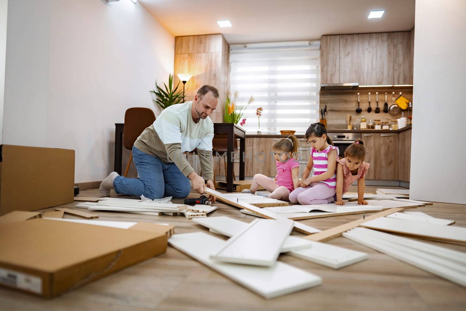 In a warm and inviting living area, a father carefully assembles furniture while surrounded by his three engaged daughters, creating a fun and collaborative home project atmosphere.