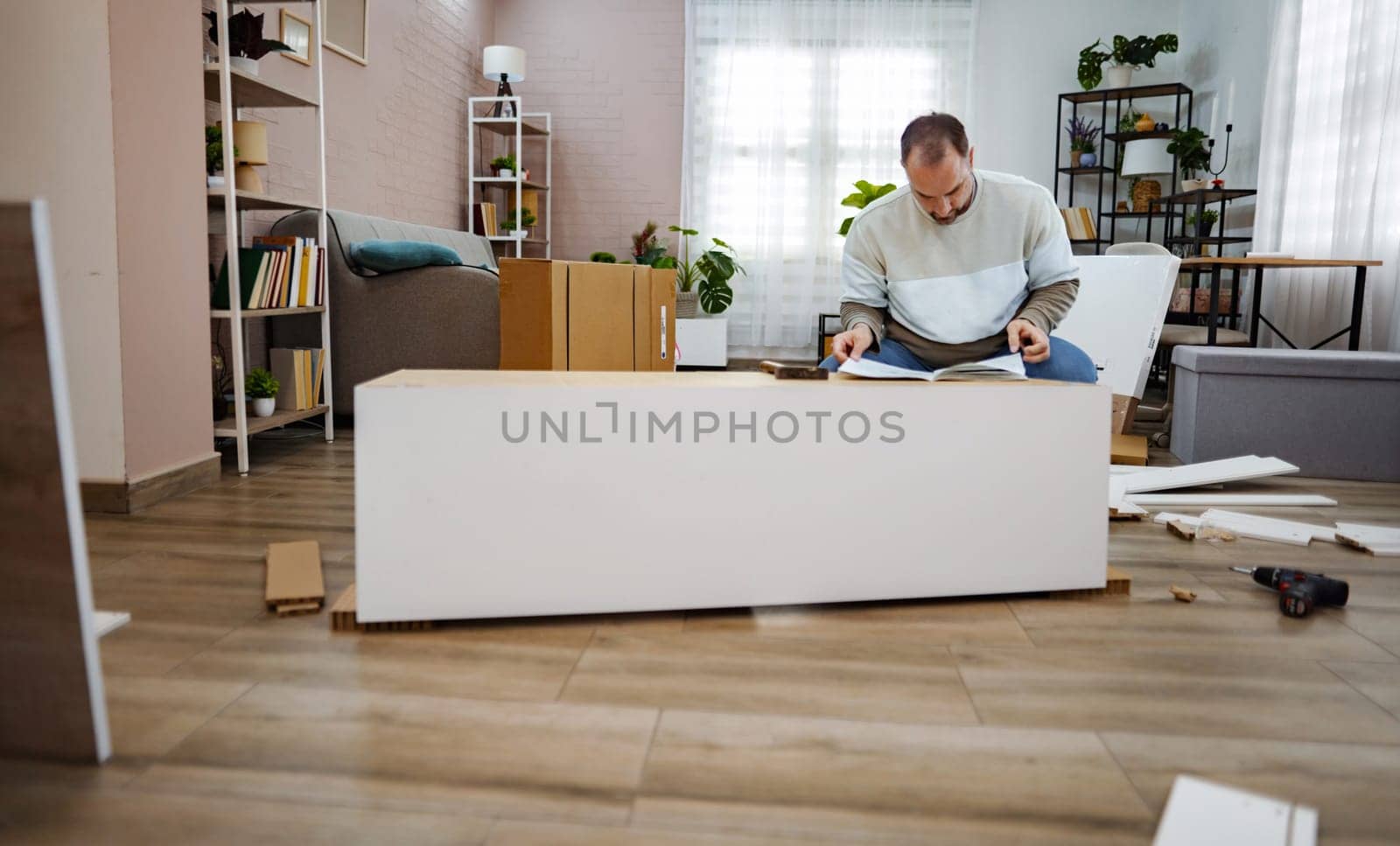 In a stylish living room filled with plants and natural light, a man intently works on assembling furniture, surrounded by boxes and tools. The cozy environment exudes a sense of creativity and focus.