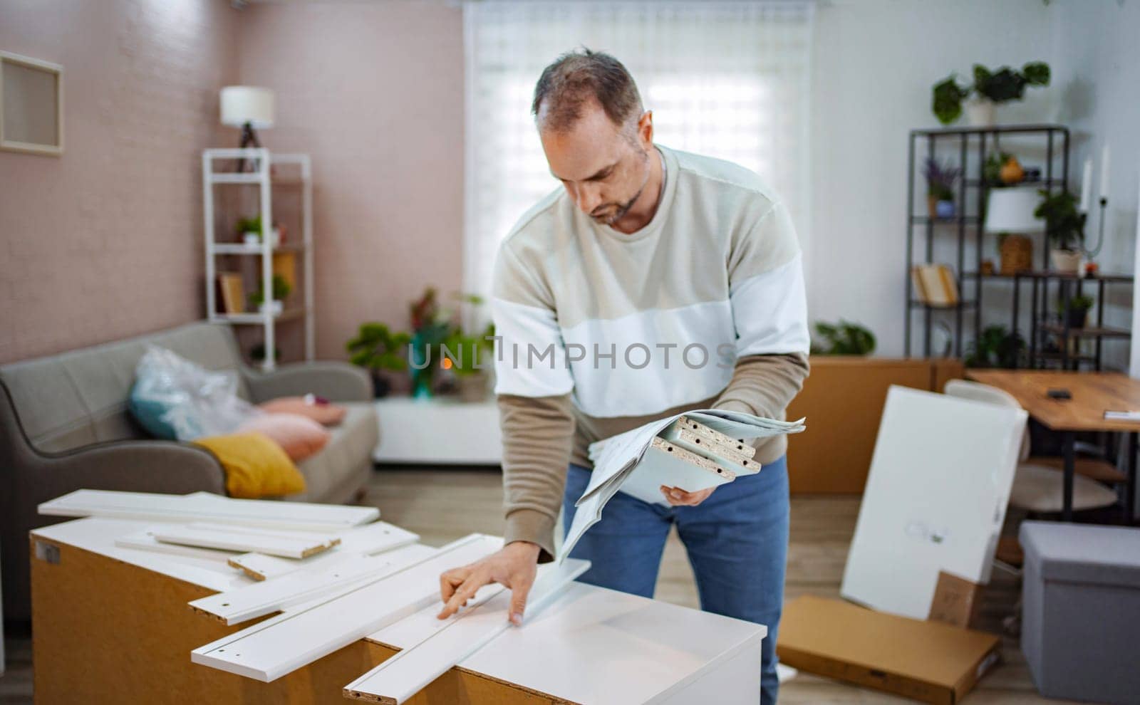 A man focuses intently on assembling furniture in a bright living room. Surrounded by parts and tools, he references instructions while creating a welcoming space filled with greenery and warmth.
