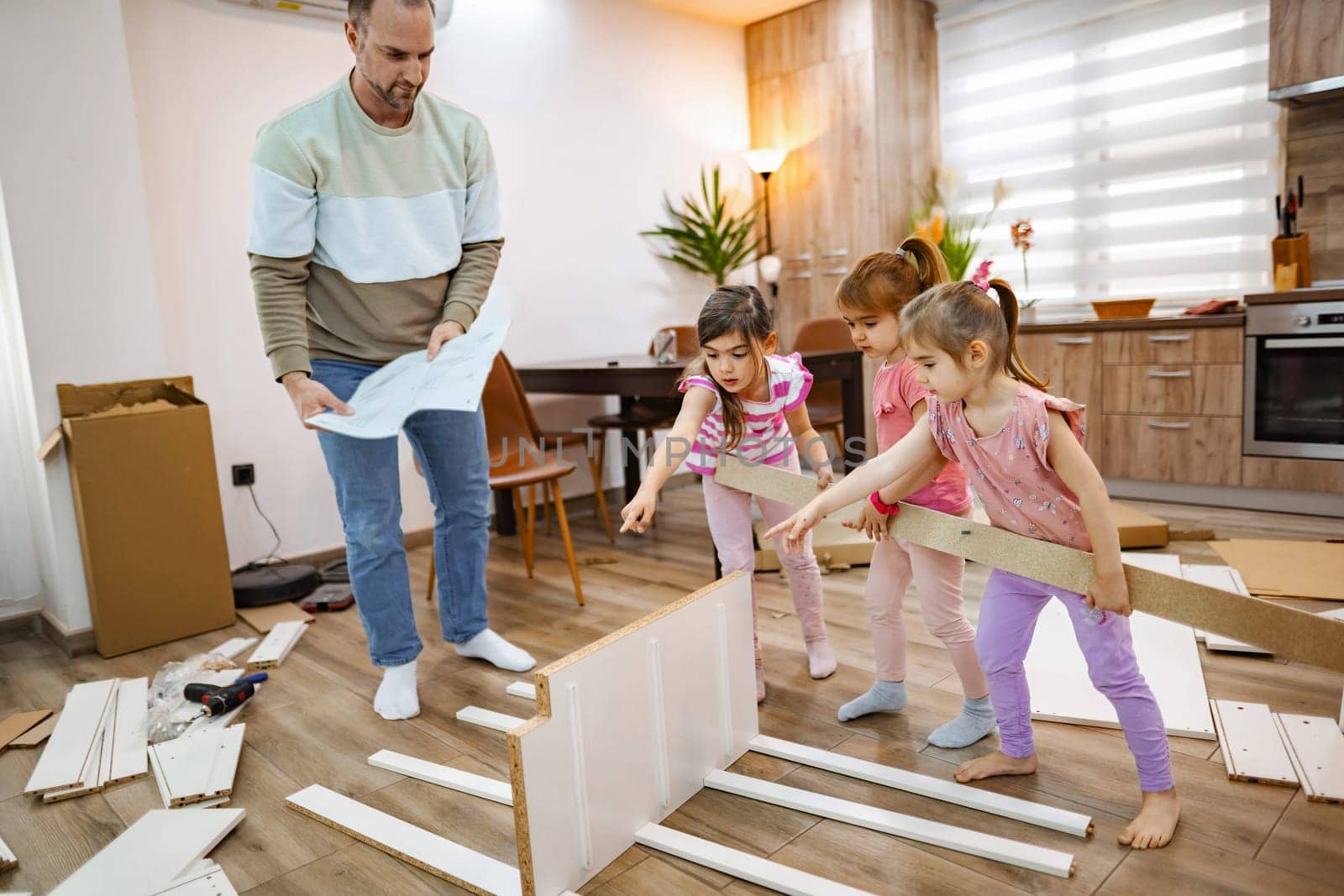 A father and his three daughters collaborate joyfully to build a new piece of furniture in their inviting living space. The room is modern, with wood accents and natural light flowing in.