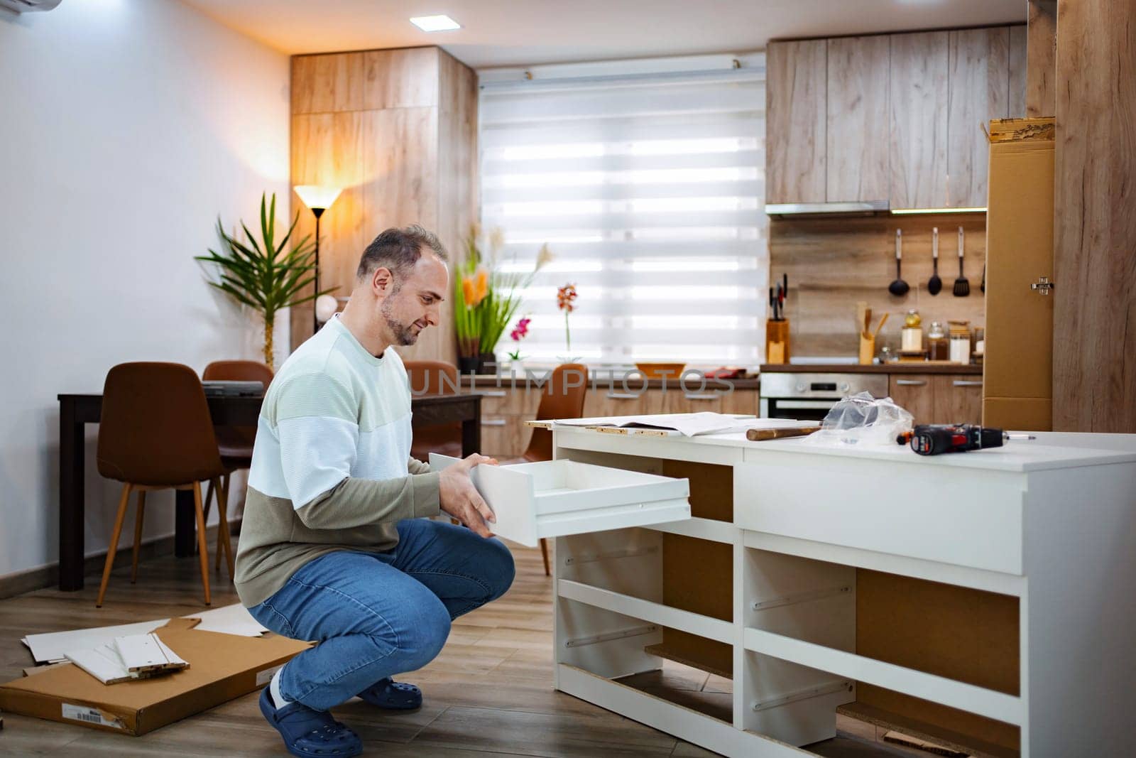 In a stylish kitchen with wooden accents, a man kneels, carefully installing a drawer into a sleek white cabinet. Sunlight filters through sheer curtains, enhancing the warm atmosphere.
