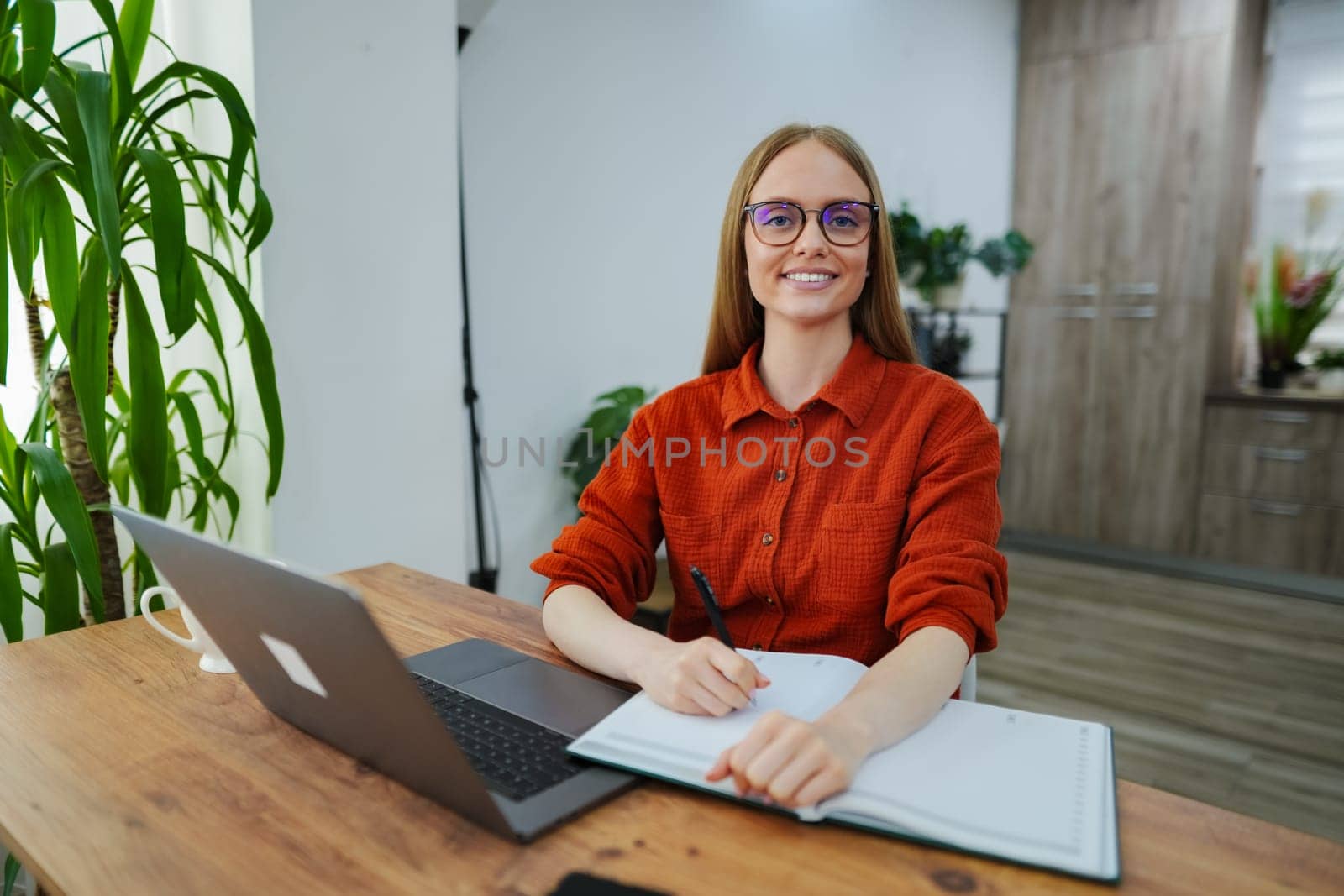 A focused woman takes notes while sitting at her desk, surrounded by greenery.