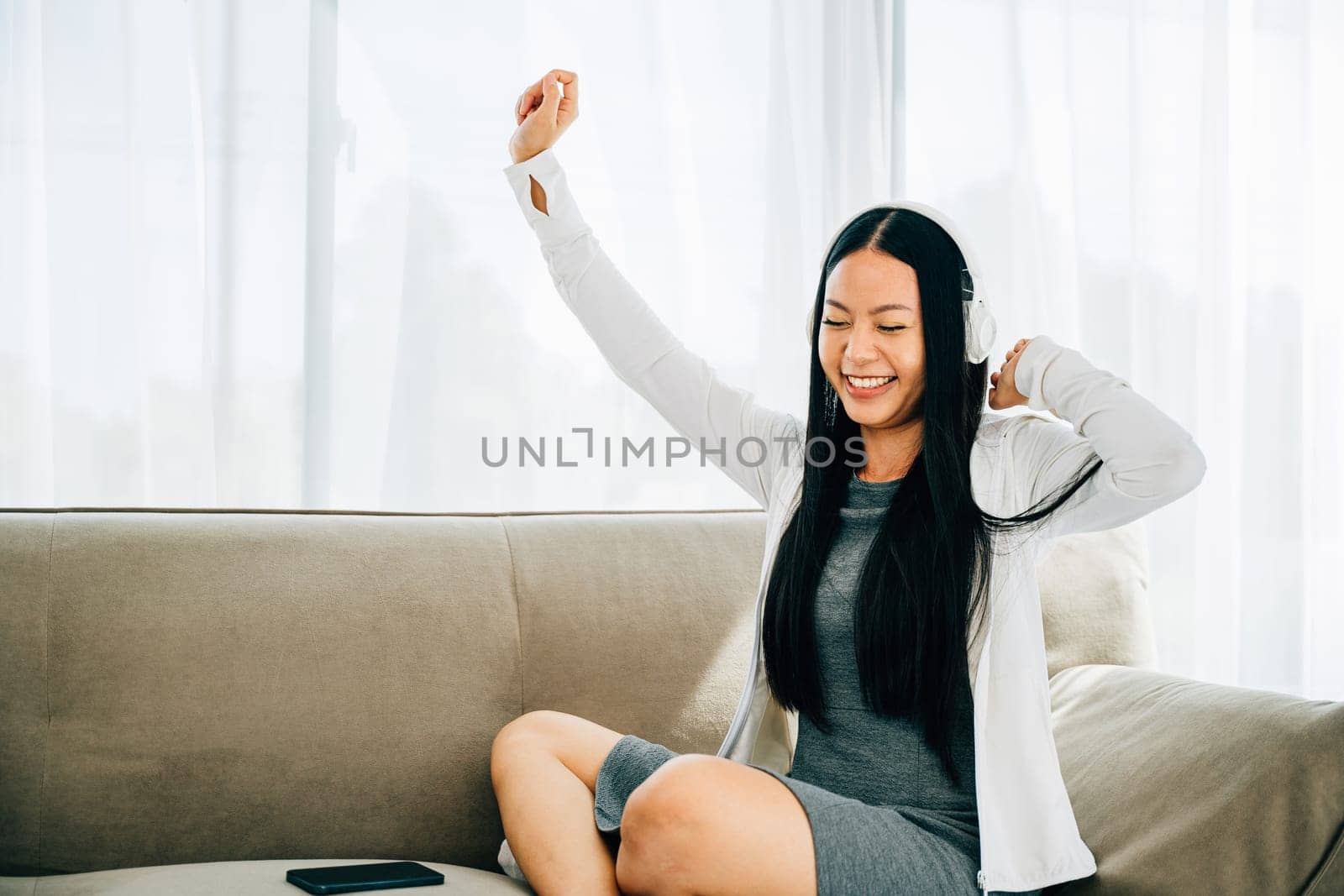Seated comfortably on the sofa a woman holds her smartphone dancing with headphones on. Immersed in music experiencing happiness and leisurely entertainment at home. Lively relaxation concept.