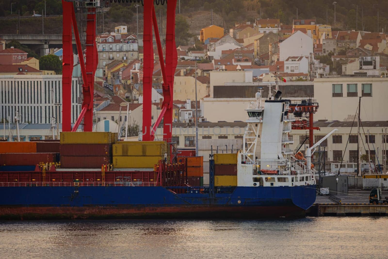 Cargo ship loaded with multicolored containers docked at Lisbon port, red cranes and city vibrant buildings on sunny evening. Dynamic residential and industrial scene by waterfront, urban skyline, loading, unloading, calm waters, commerce, industry