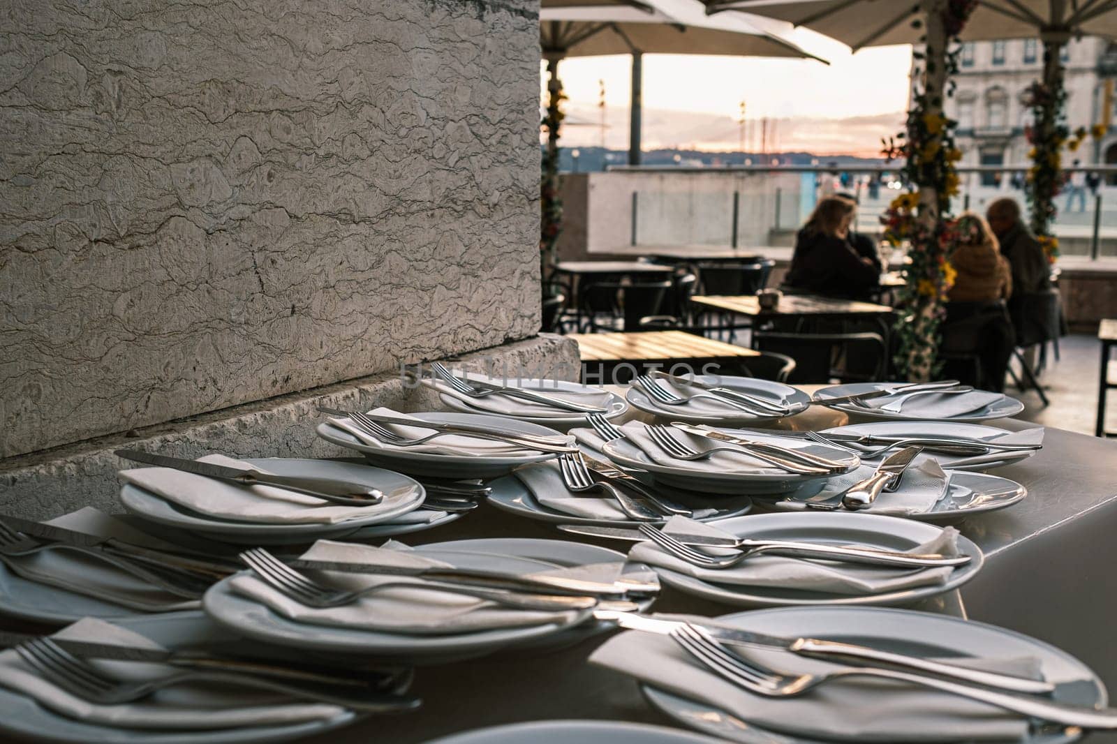 Plates neatly arranged with forks, knives, and napkins on stone countertop at outdoor restaurant in selective focus. Blurred background with European city square at sunset. Relaxed dining, people seated, umbrellas, travel destination, summer vacation