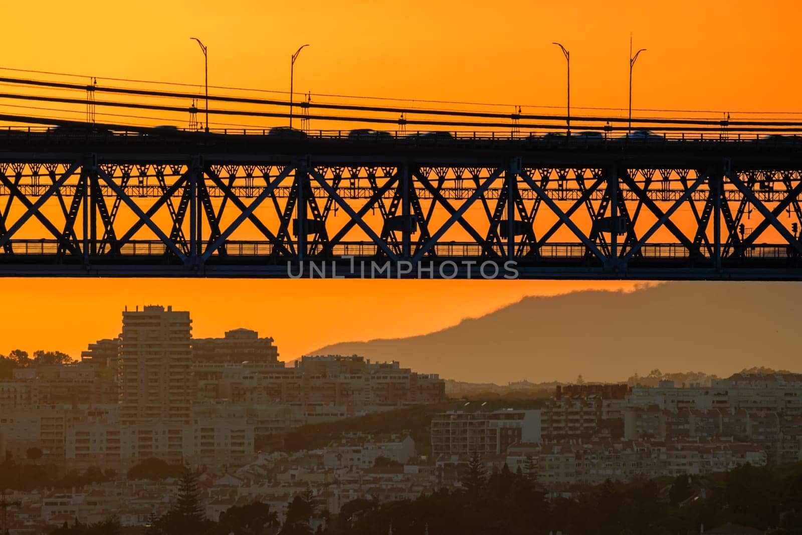 Dramatic close view of section of iconic 25 de Abril bridge in silhouette against vibrant orange sunset sky, Lisbon, Portugal cityscape and distant hills below in summer sundown haze in background, vehicles run on bridge in motion blur