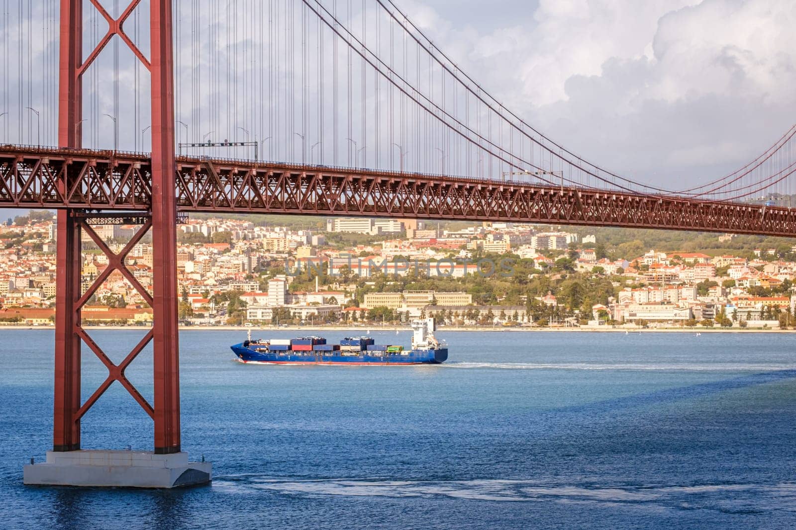 Colorful landscape with blue container ship on Tagus river under iconic 25 de Abril or 25th April bridge, Lisbon, Portugal. Famous ironwork of red suspension bridge, vibrant cityscape, urban architecture, maritime transport, sunny day, cloudy sky