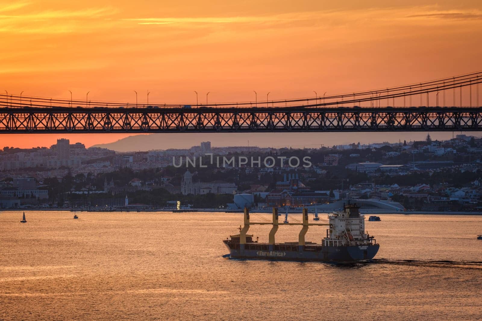 Cargo ship sail on Tagus river under iconic 25 de Abril bridge in silhouette, Lisbon, Portugal at sunset. City skyline, glowing orange sky, dramatic maritime and urban scene, European port, summer haze over horizon, tiny sailboats on calm waters.