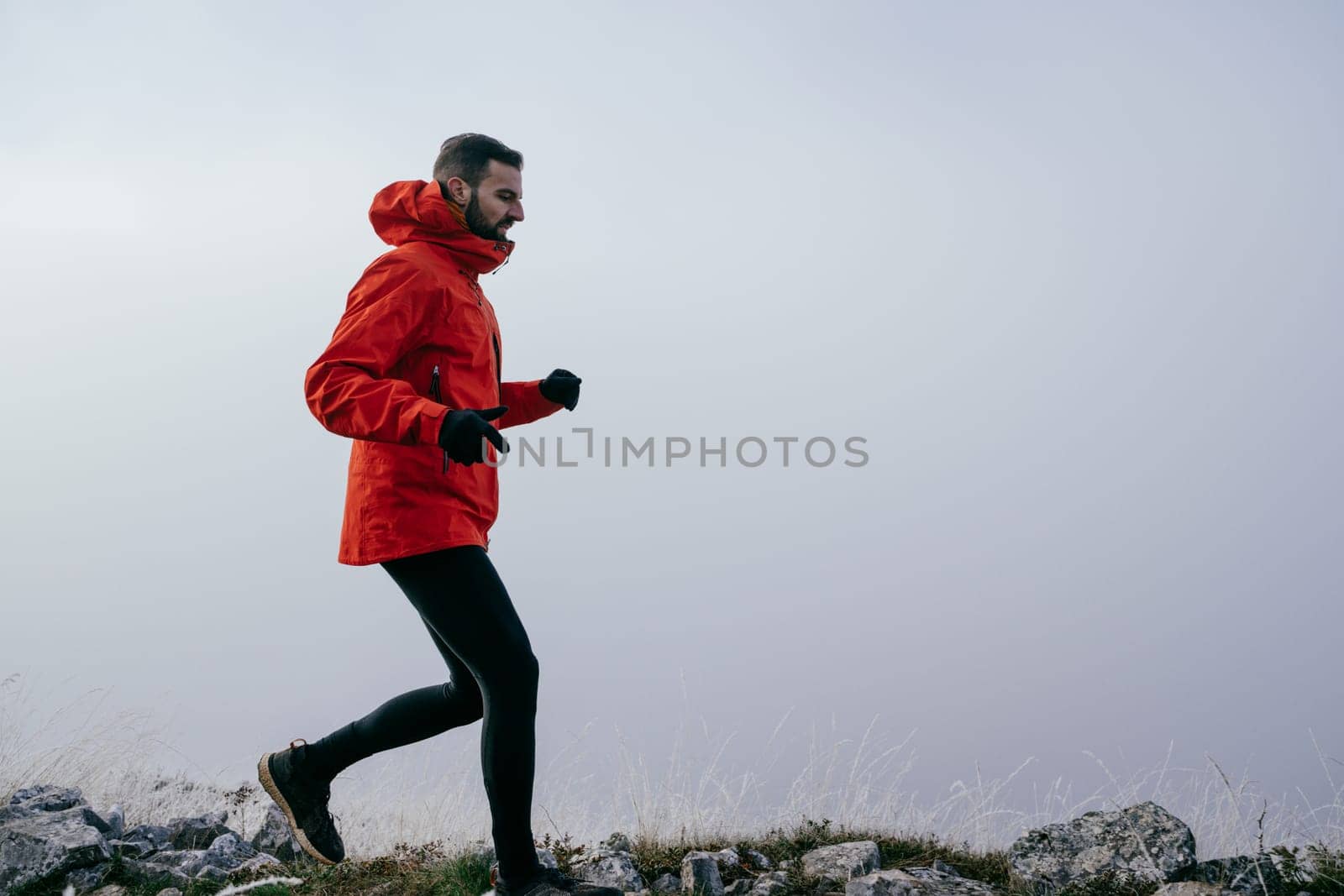 A man in athletic wear climbs a rocky hill in foggy weather. Dressed in a red jacket and black leggings, he is captured mid stride in a determined motion against a pale, misty background.