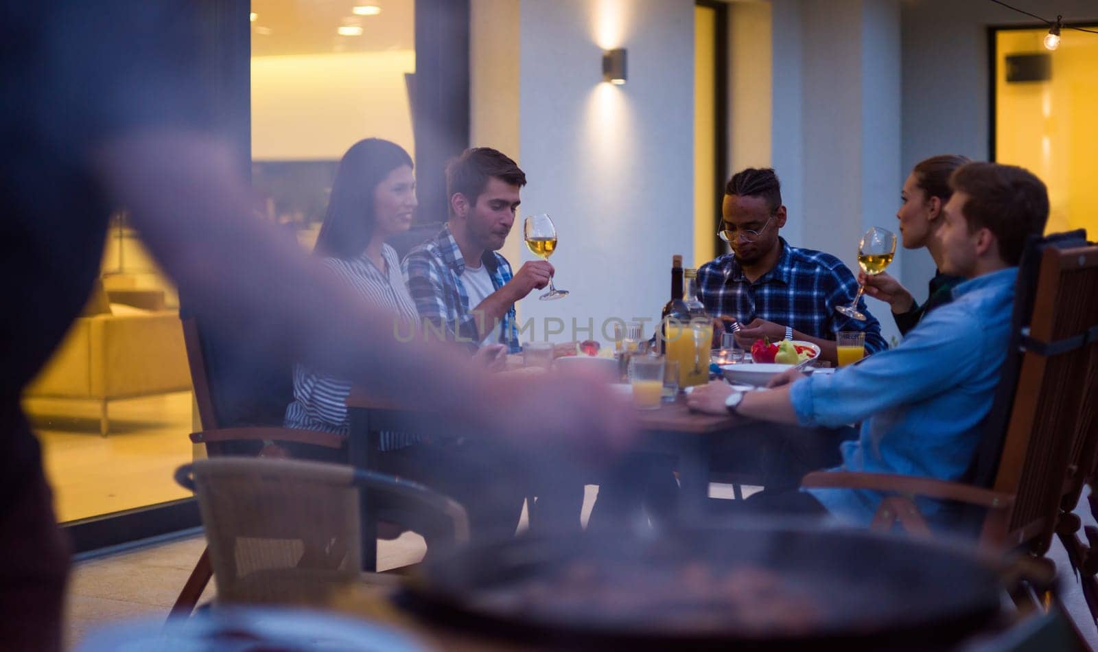 A group of young diverse people having dinner on the terrace of a modern house in the evening. Fun for friends and family. Celebration of holidays, weddings with barbecue.