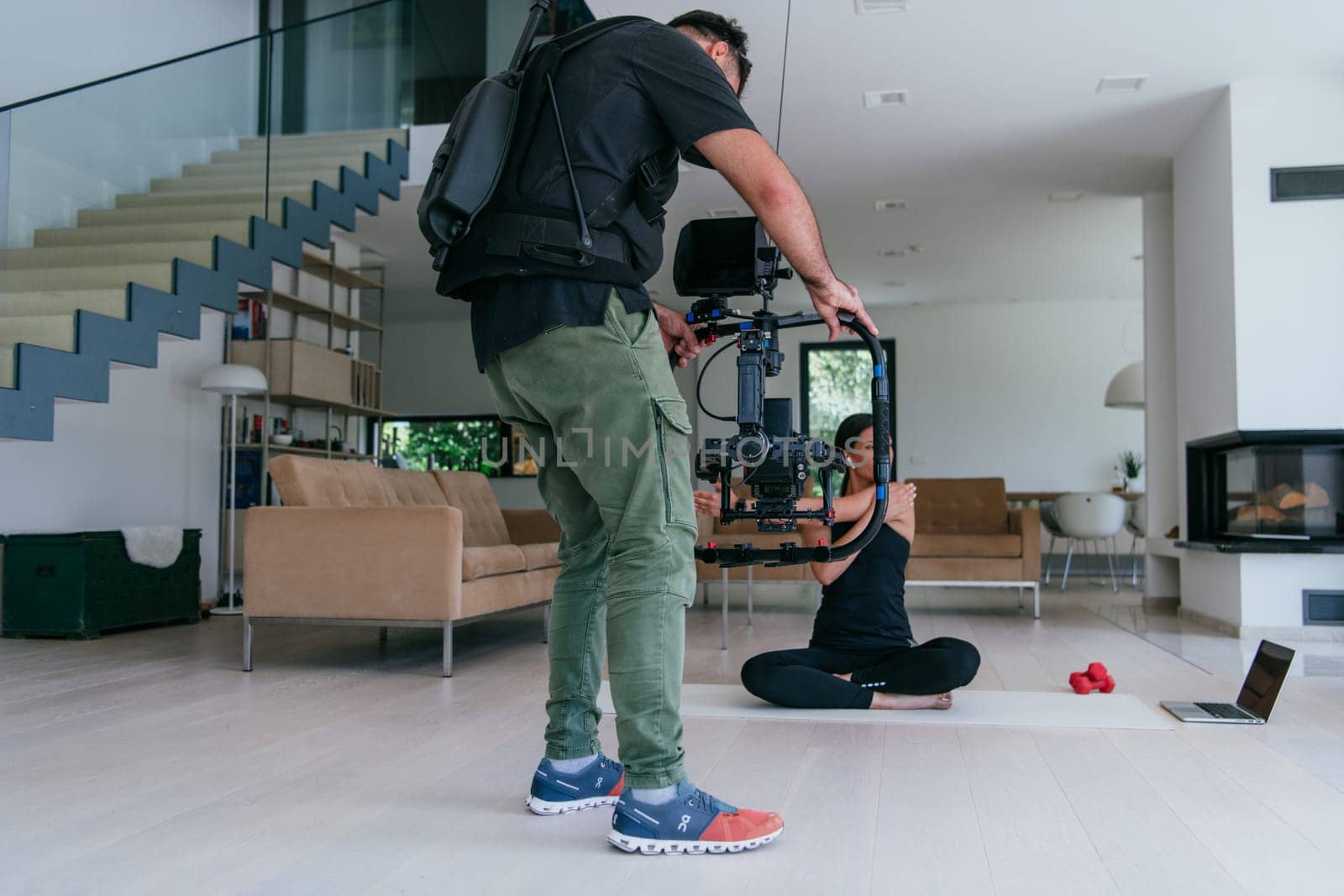 Behind the scene. Photo of a cameraman recording a woman doing yoga exercises at an online training while using and watching a laptop