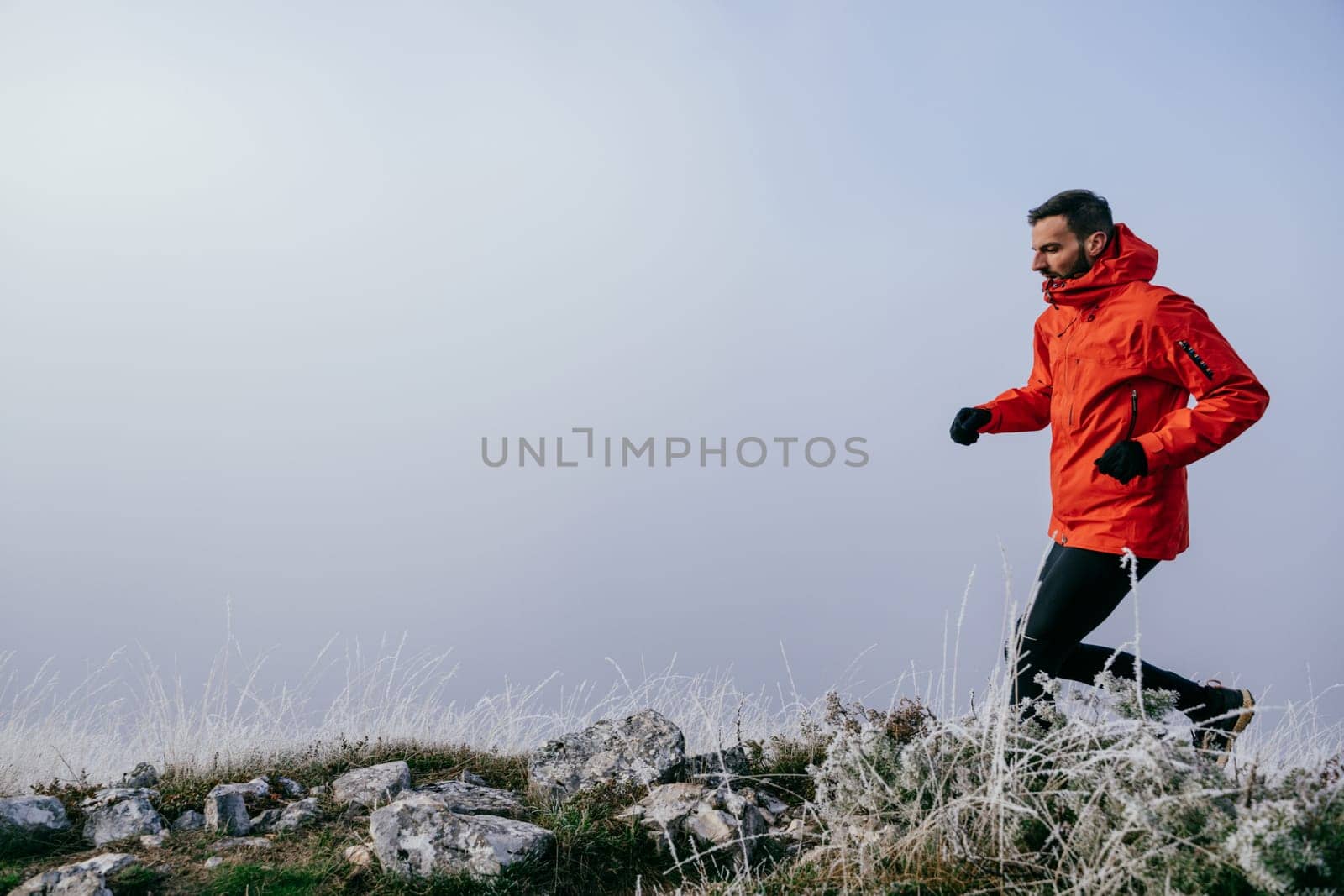 A man in athletic wear climbs a rocky hill in foggy weather. Dressed in a red jacket and black leggings, he is captured mid stride in a determined motion against a pale, misty background.