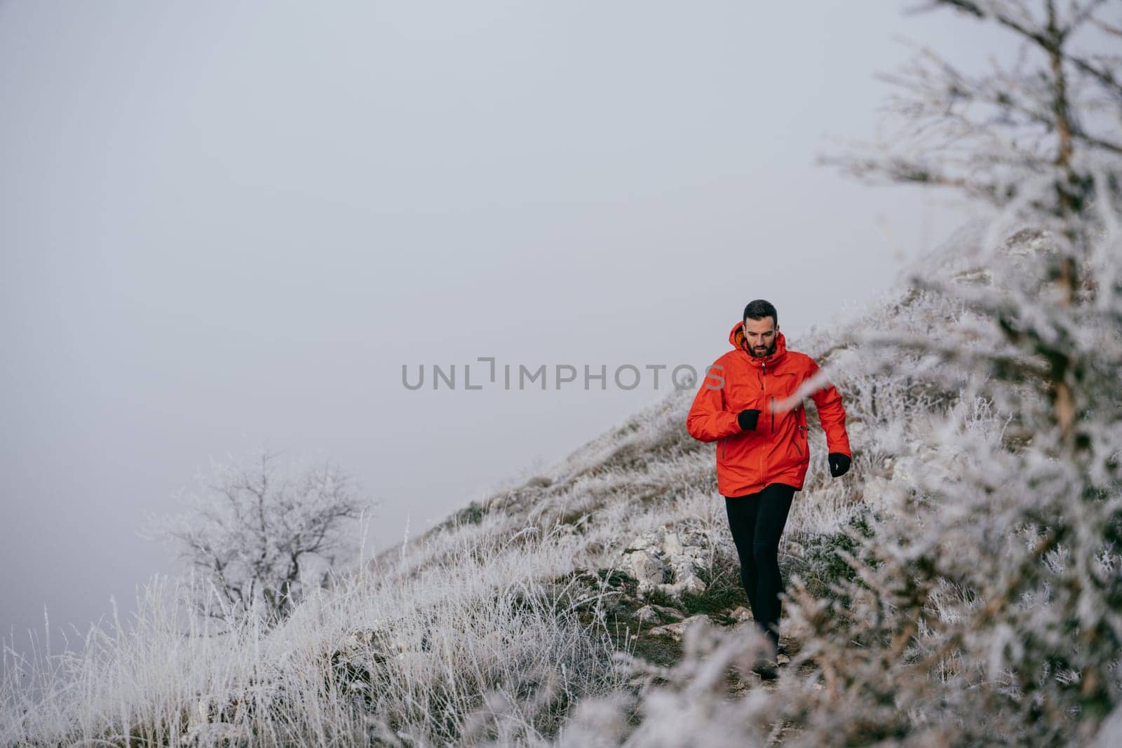 A man in athletic wear climbs a rocky hill in foggy weather. Dressed in a red jacket and black leggings, he is captured mid stride in a determined motion against a pale, misty background.