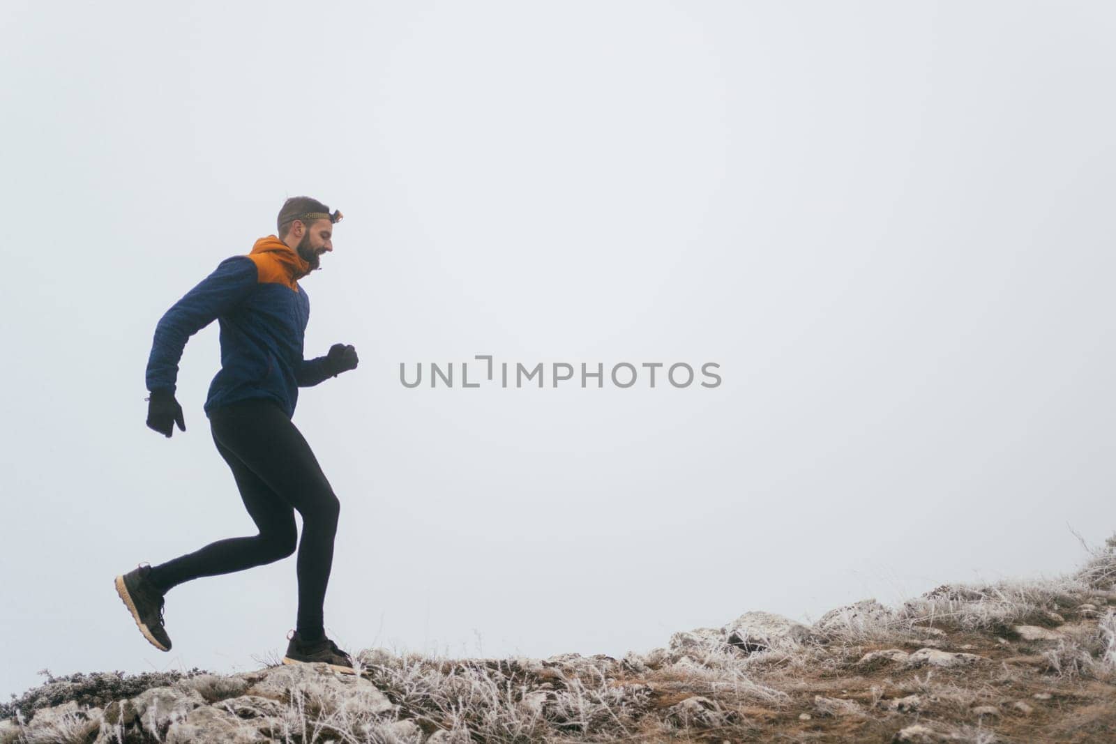 A man in athletic wear climbs a rocky hill in foggy weather. Dressed in a blue jacket and black leggings, he is captured mid-stride in a determined motion against a pale, misty background.