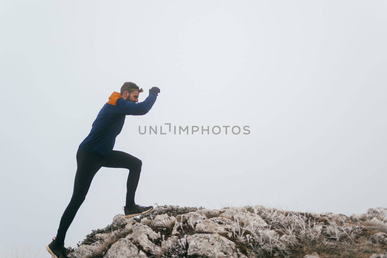 A man in athletic wear climbs a rocky hill in foggy weather. Dressed in a blue jacket and black leggings, he is captured mid-stride in a determined motion against a pale, misty background.