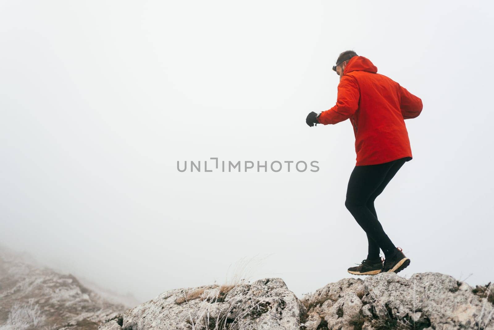 A man in athletic wear climbs a rocky hill in foggy weather. Dressed in a red jacket and black leggings, he is captured mid stride in a determined motion against a pale, misty background.