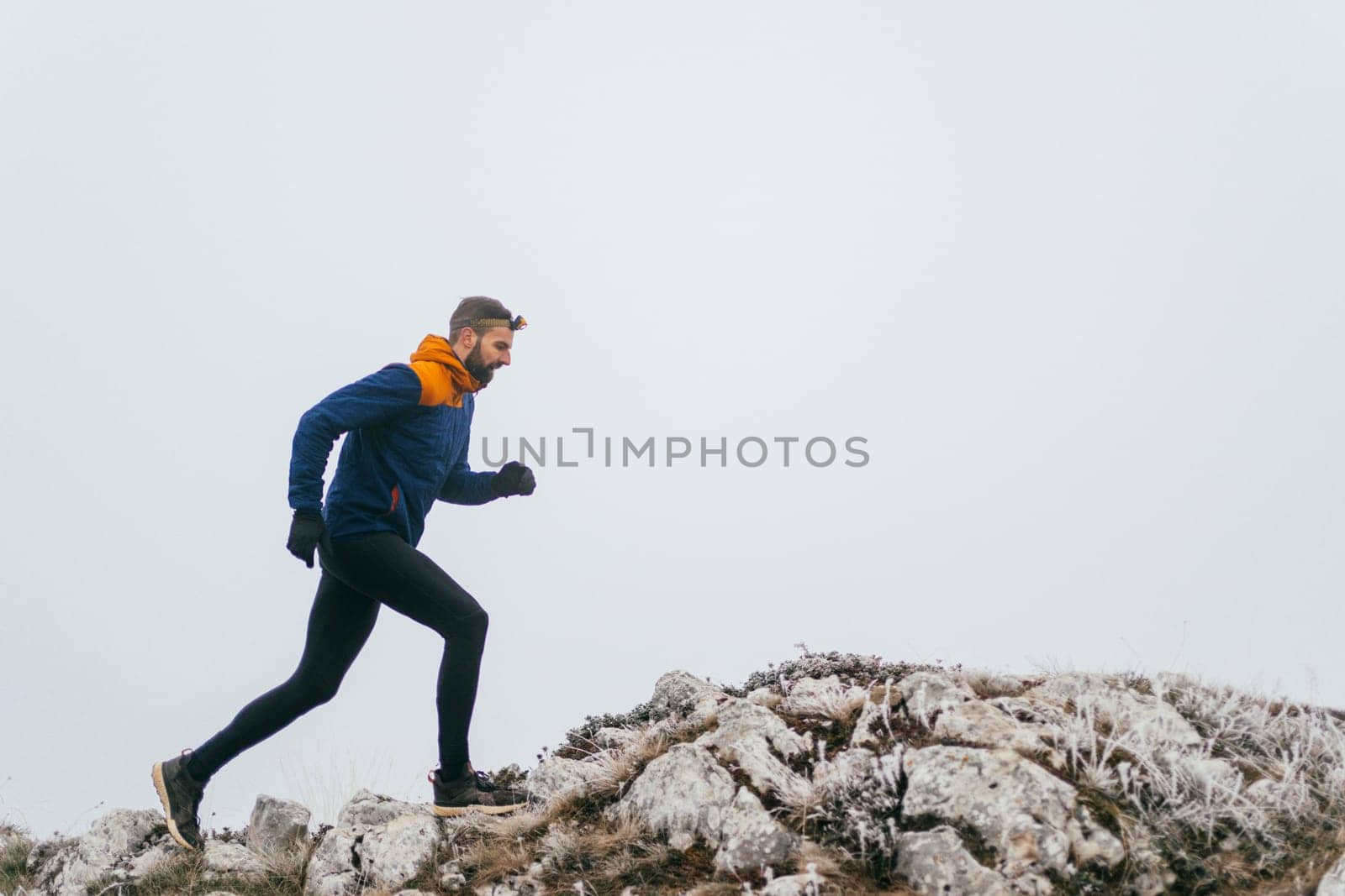 A man in athletic wear climbs a rocky hill in foggy weather. Dressed in a blue jacket and black leggings, he is captured mid-stride in a determined motion against a pale, misty background.