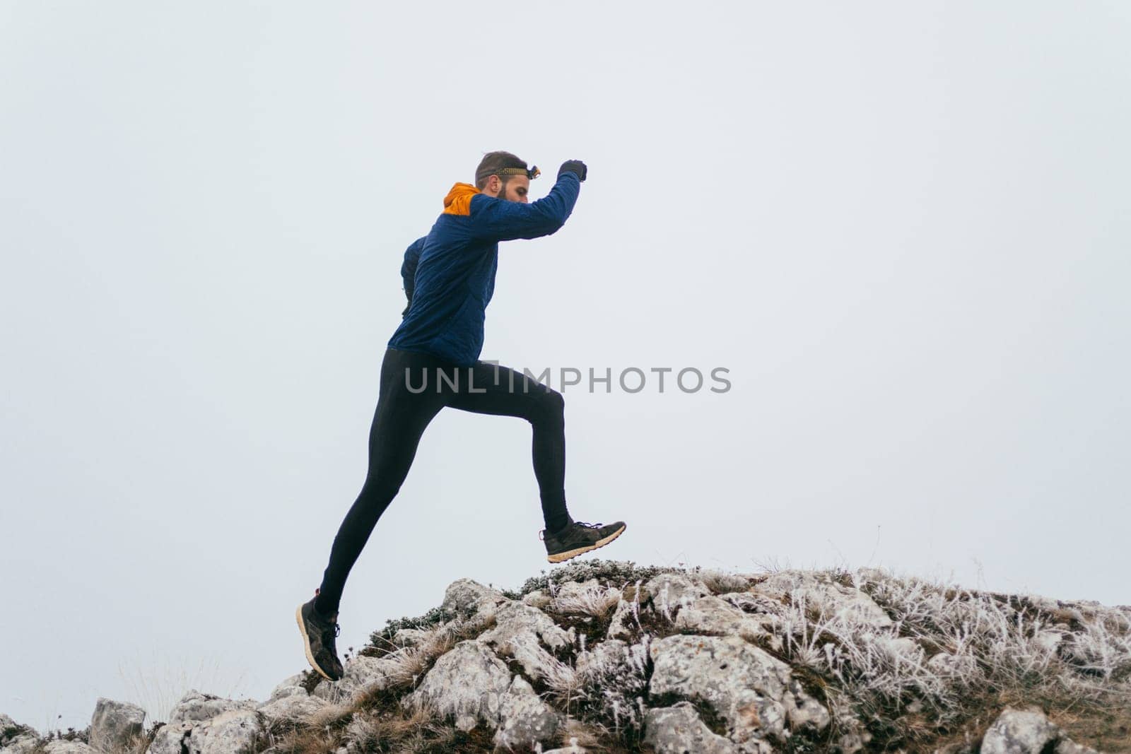 A man in athletic wear climbs a rocky hill in foggy weather. Dressed in a blue jacket and black leggings, he is captured mid-stride in a determined motion against a pale, misty background.