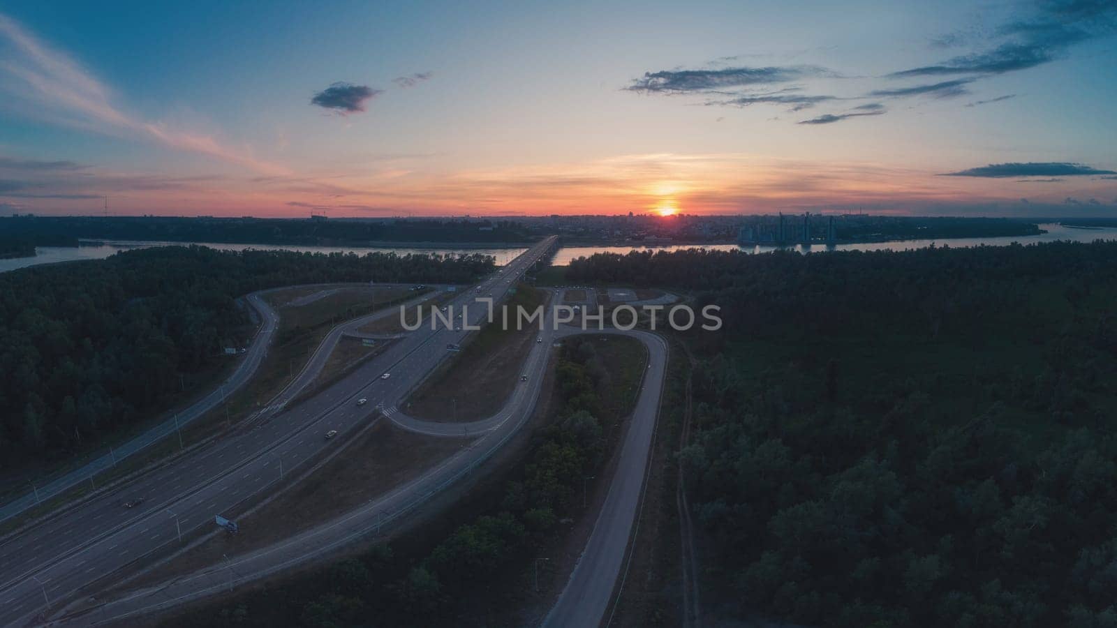 Drone photo of a highway bridge stretching over a river toward a Barnaul city skyline, captured at colorful sunset with clear sky.