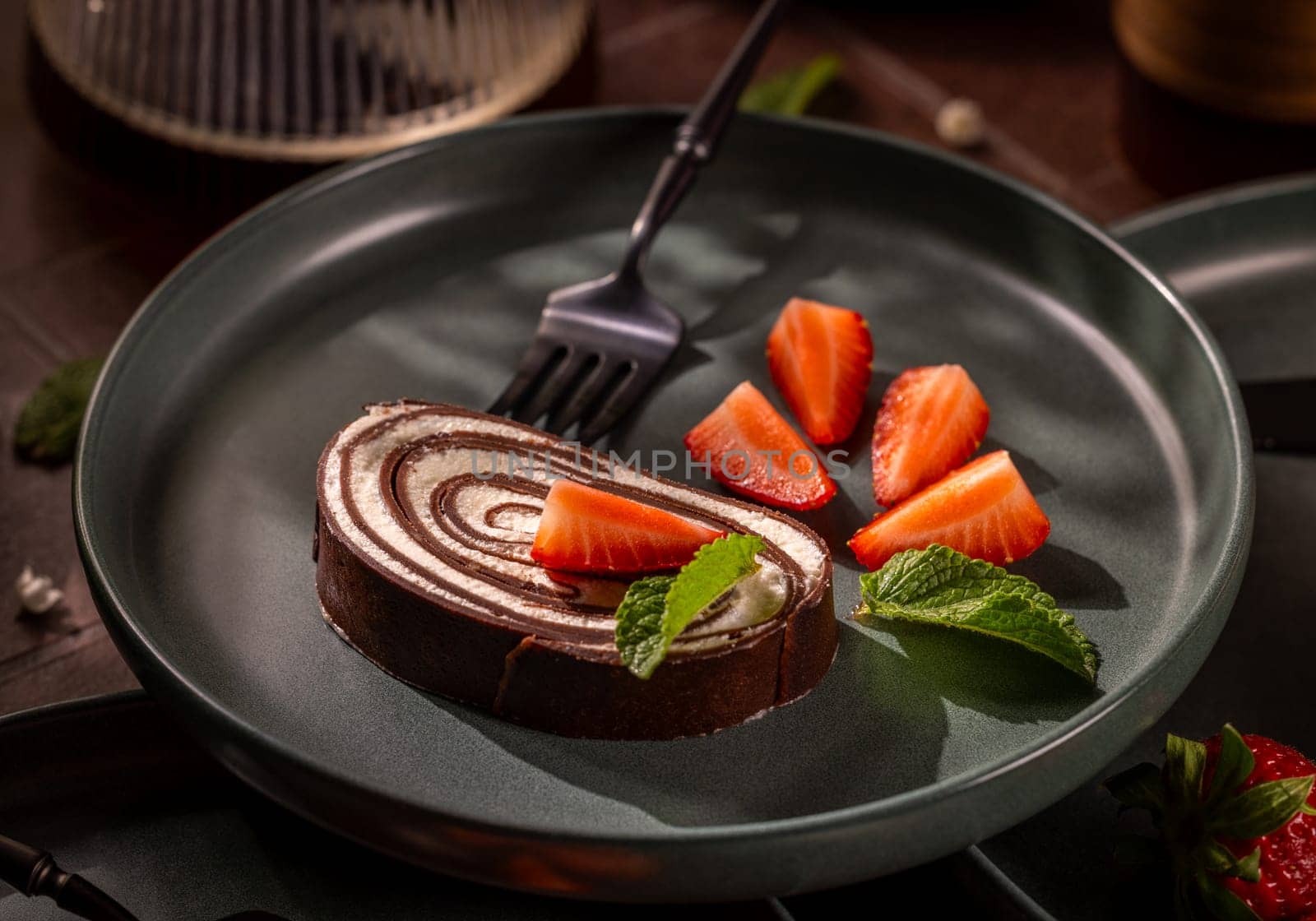 Delicious chocolate swiss roll served with fresh strawberry slices and mint leaves on a dark plate, creating an elegant dessert presentation