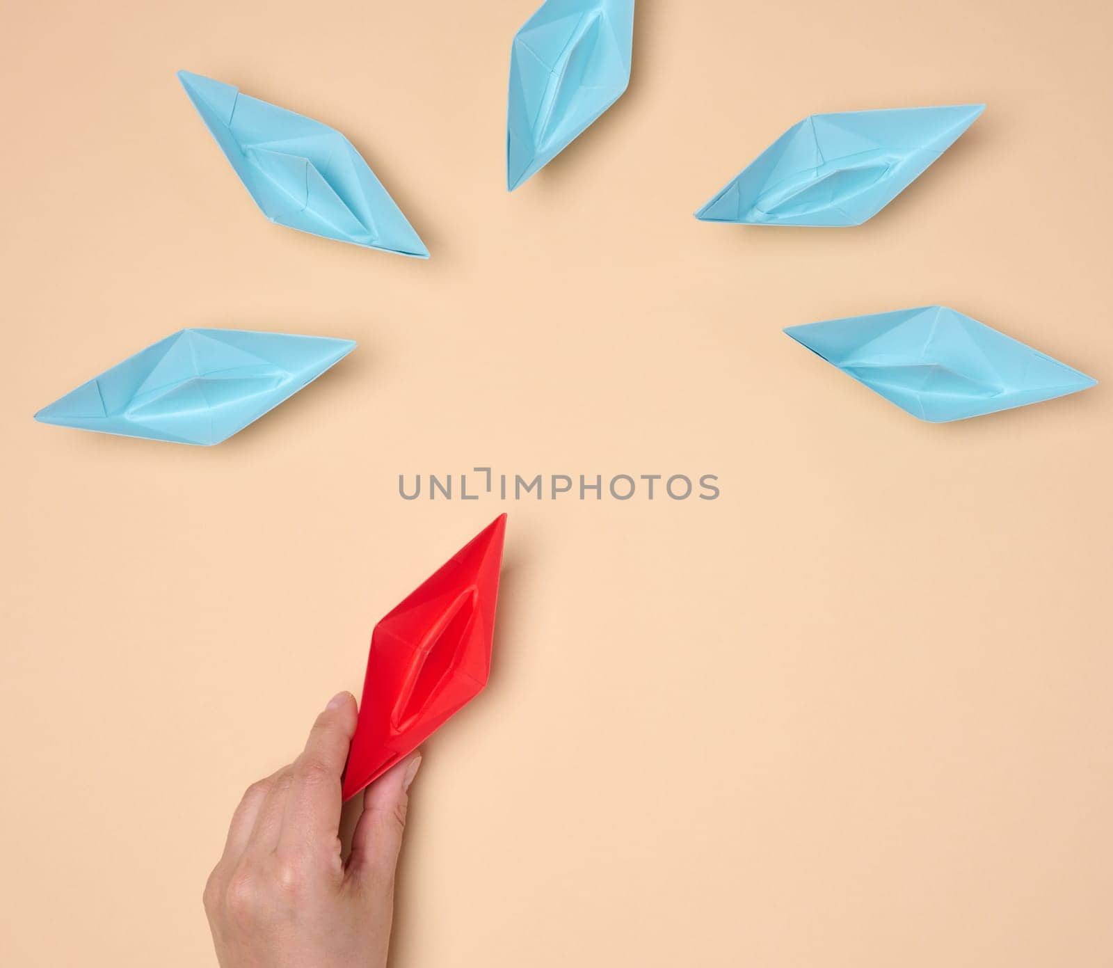 A hand holds a red origami paper boat next to a group of blue ones, symbolizing choice, uniqueness or a new idea, on a neutral background.