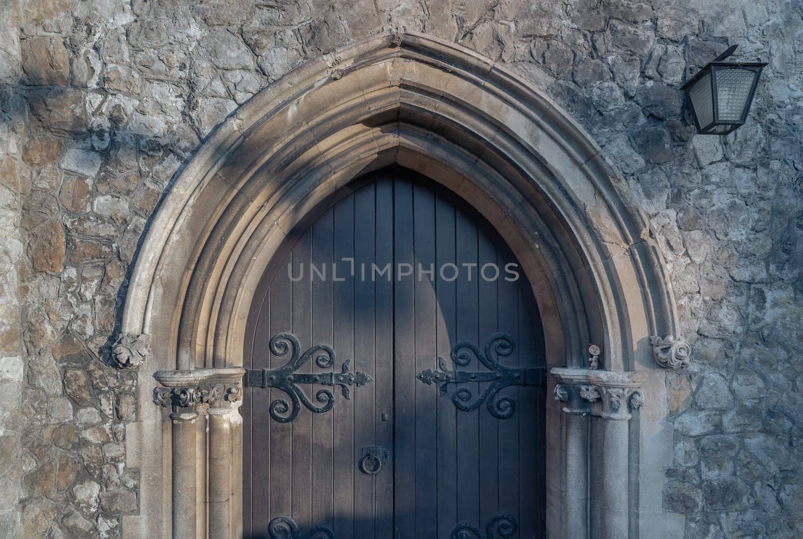 Royal Tunbridge Wells, UK - Apr 29, 2025 - Detail exterior of Old gothic medieval church wooden door and sandstone arched with decorative wrought iron hinge. Sunlight shines through on the surface of Ornamented wooden gate, use it as your Wallpaper, Poster and Space for text, Selective focus.