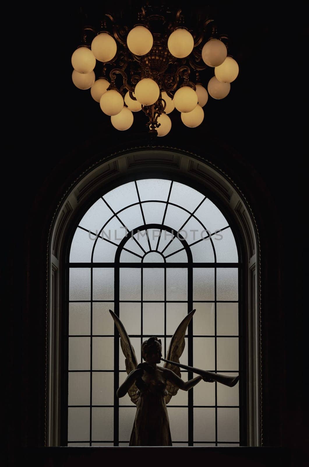 London, UK - May 07, 2025 - Silhouette view of Outstanding Angel of Gaiety statue (Spirit of Gaiety) in front of Large semi oval glass window and Hanging lantern from the ceiling on top at the Victoria and Albert Museum. Abstract interior design, use it as your Wallpaper, Poster and Space for text, Selective focus.