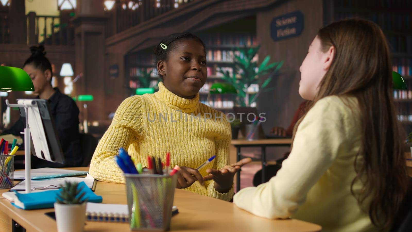 Happy two girls emphasizing their friendship with an embrace at the library, taking a break from study session and chatting about something. Classmates and friends having fun together. Camera A.