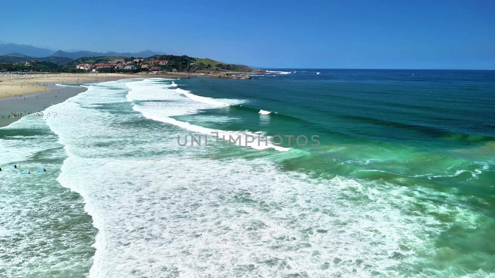 An aerial view shows a serene beach with clear turquoise water, gentle waves, and a vibrant surrounding landscape