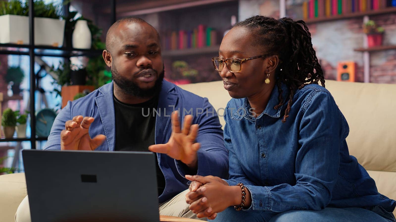 Black young couple attending an online video call connection at home, sitting on the couch and talking to their friends. Man and woman waving on a video conference, lifestyle. Camera B.