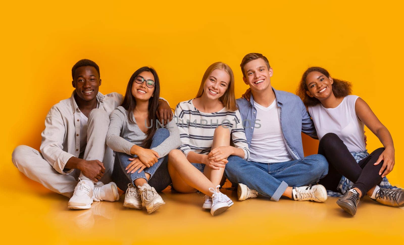 Multiracial joyful students sitting on floor, embracing and having fun over yellow studio background, students lifestyle concept, panorama