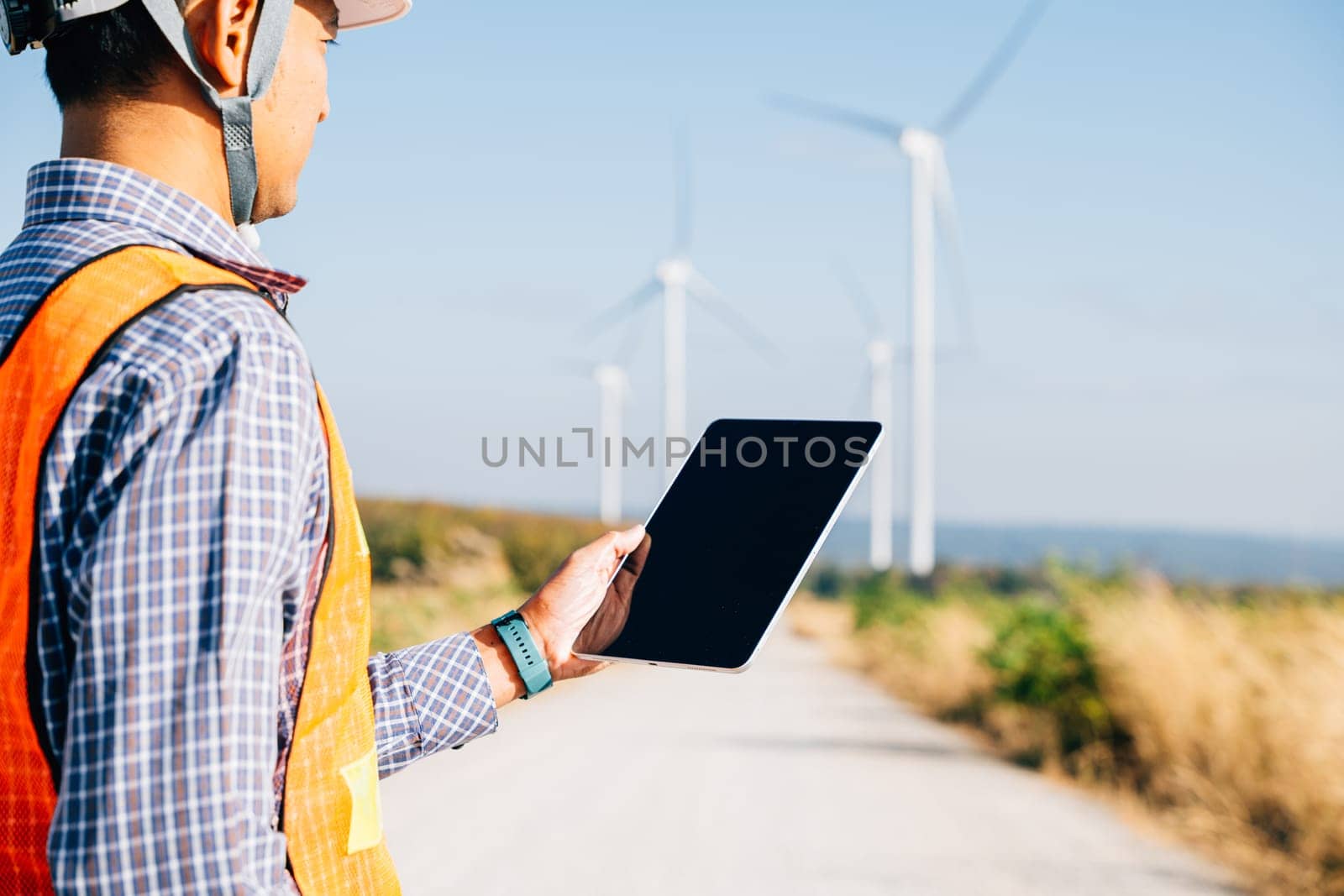 Man in hardhat using tablet at windmills. Engineer assures clean energy generation. Expertise in turbine efficiency and global electricity control portrayed confidently.