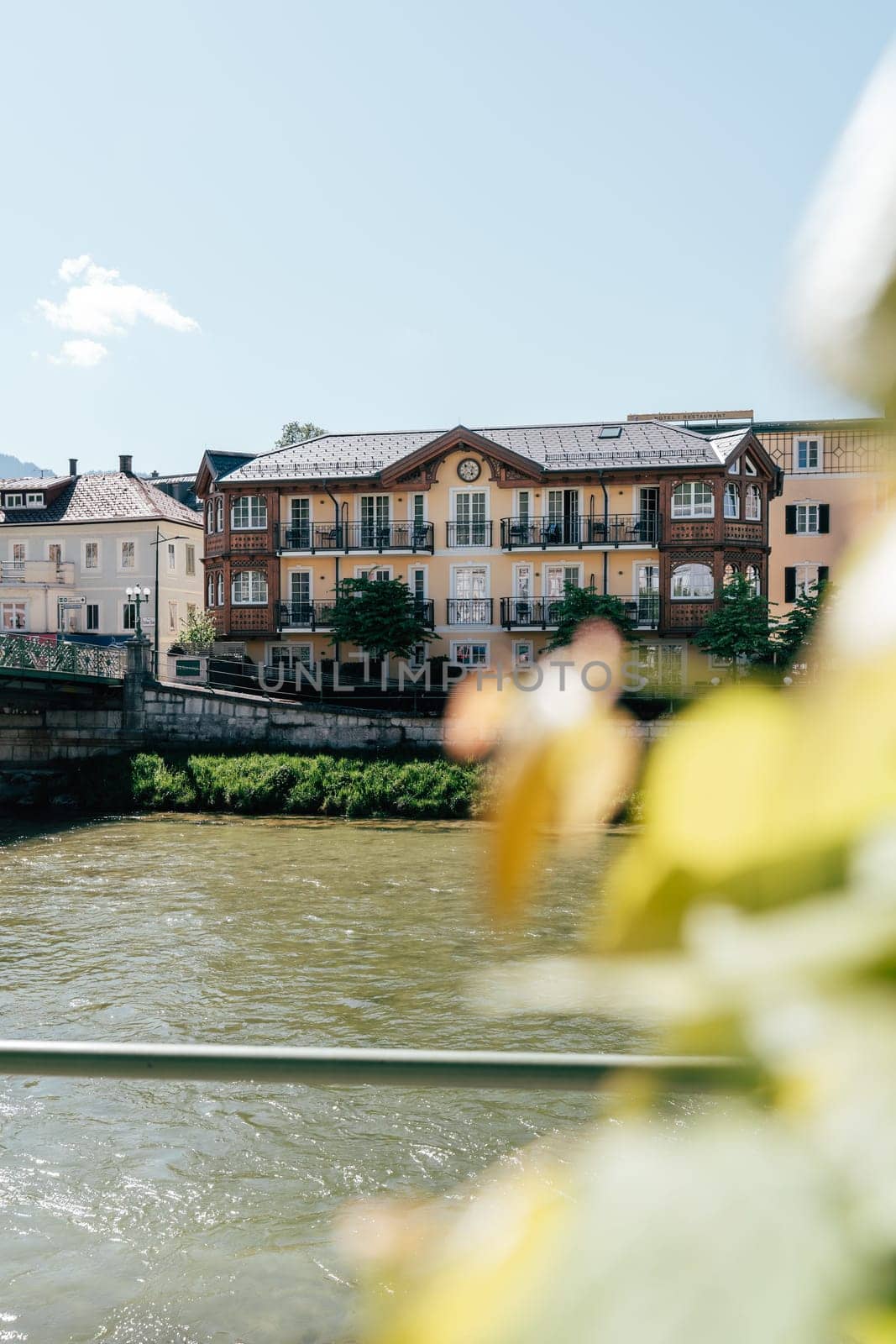 A vibrant building stands along the riverbank in Bad Ischl under a clear blue sky. Lush greenery frames the scene, highlighting the charm of this picturesque location.