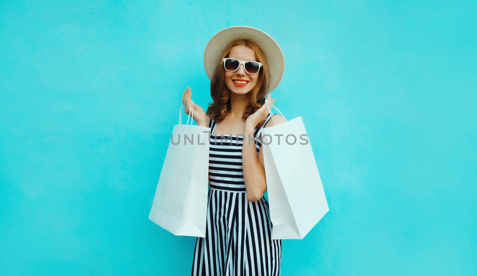Fashion, purchase. Portrait of stylish beautiful happy young woman posing with white shopping bags, girl wear summer hat, dress standing on bright blue background