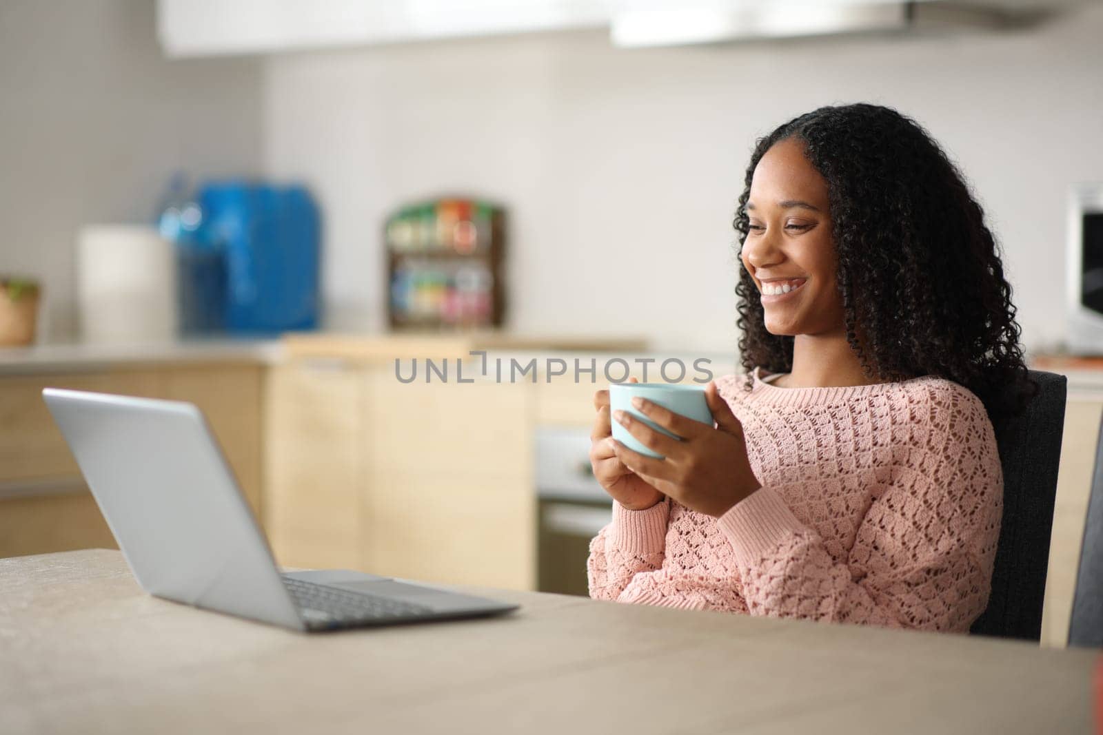 Happy black woman watching media on laptop drinking coffee in a kitchen at home