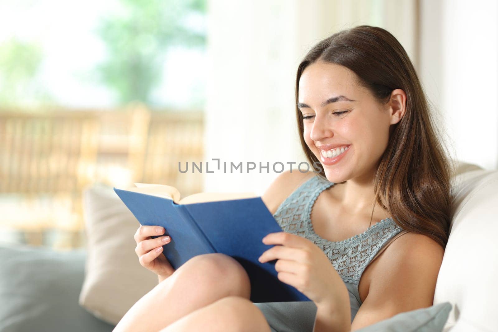 Happy woman reading a paper book sitting on a couch in a house interior