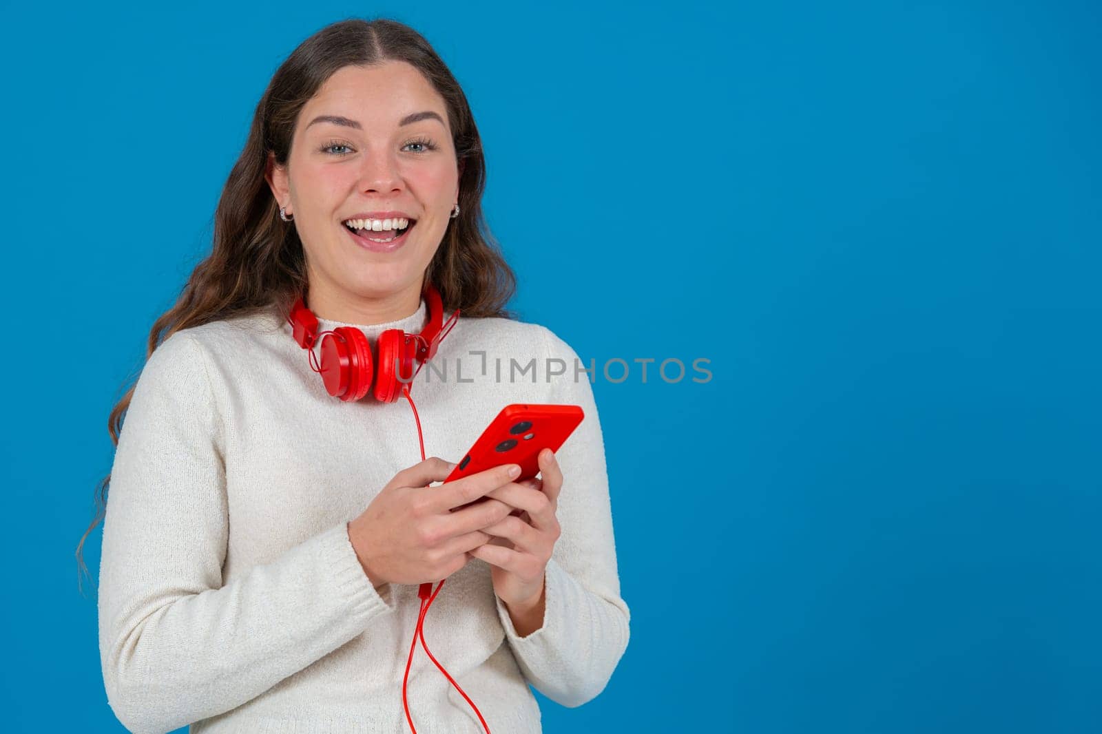 Young woman wearing red headphones around her neck, smiling while looking at her smartphone. She appears happy and excited,possibly reacting to a message or content.looking at camera, Blue background.