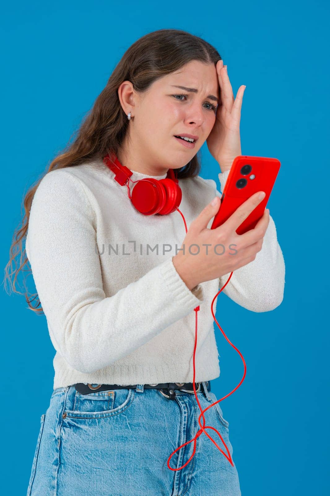 Caucasian woman looks shocked while reading bad news on her smartphone. She wears headphones and a white sweater, reacting with concern against a solid blue background.