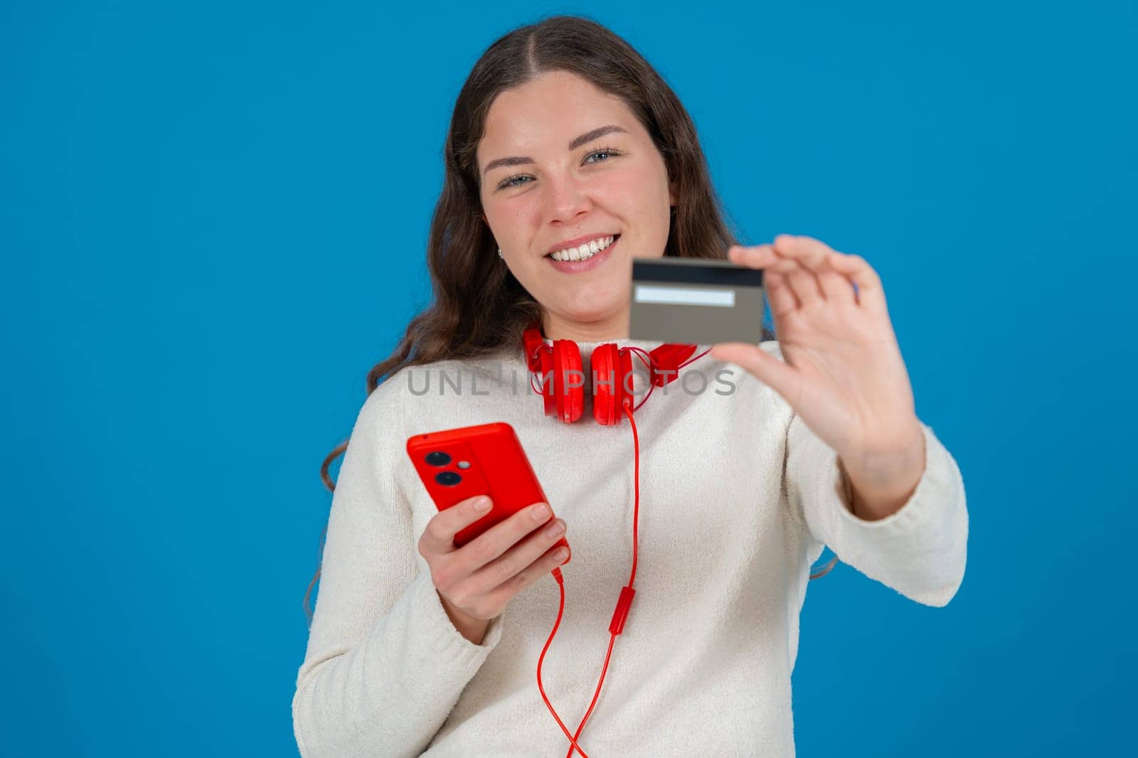 Happy young woman in a white sweater and red headphones smiling while showing a credit card to the camera and holding a red smartphone, against a blue background, symbolizing online shopping.
