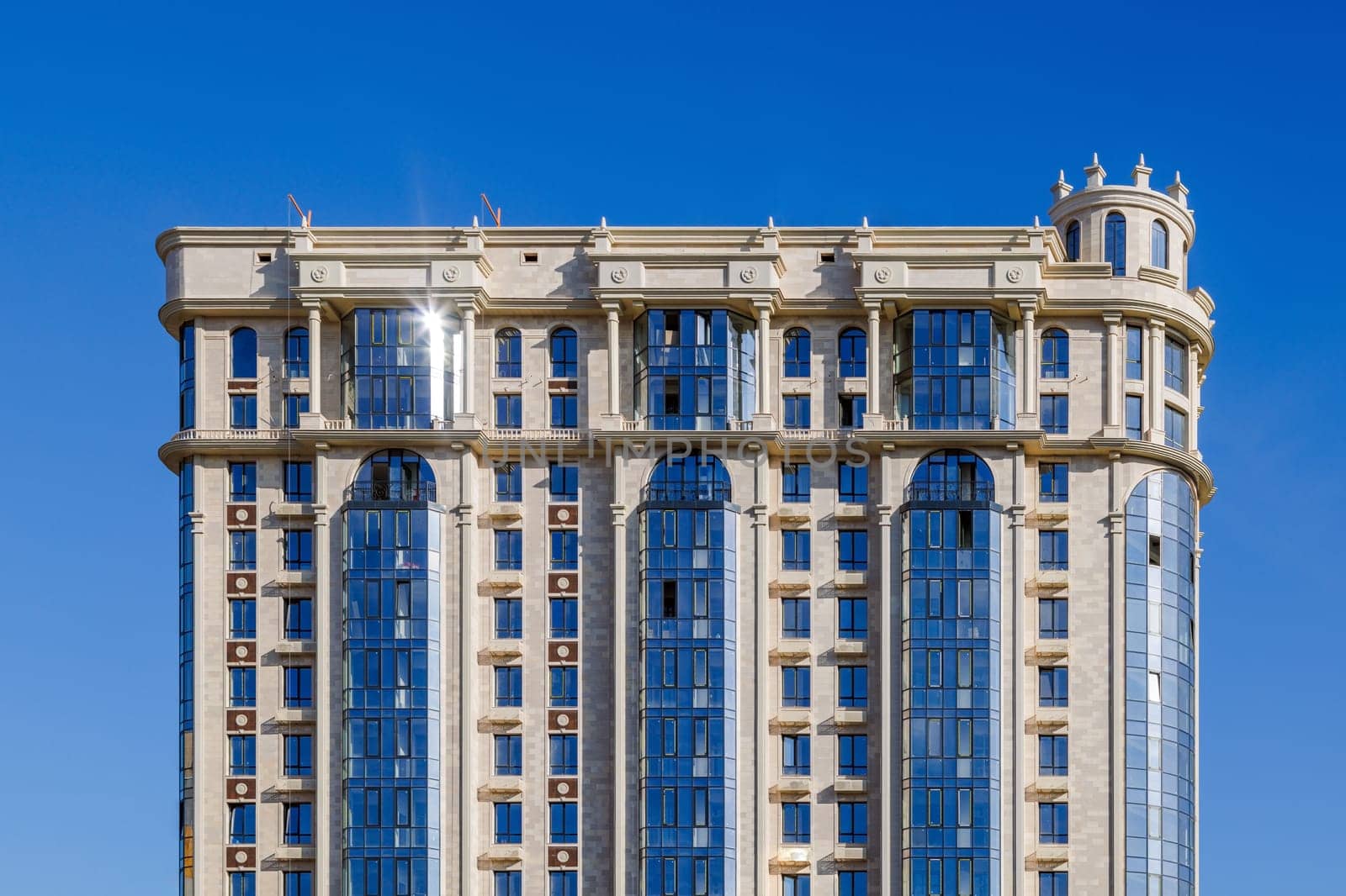 A neoclassical high-rise in Kyrgyzstan featuring a beige stone facade, arched windows, vertical columns, and reflective blue glass panels under a clear sky, blending traditional and modern architectural elements.
