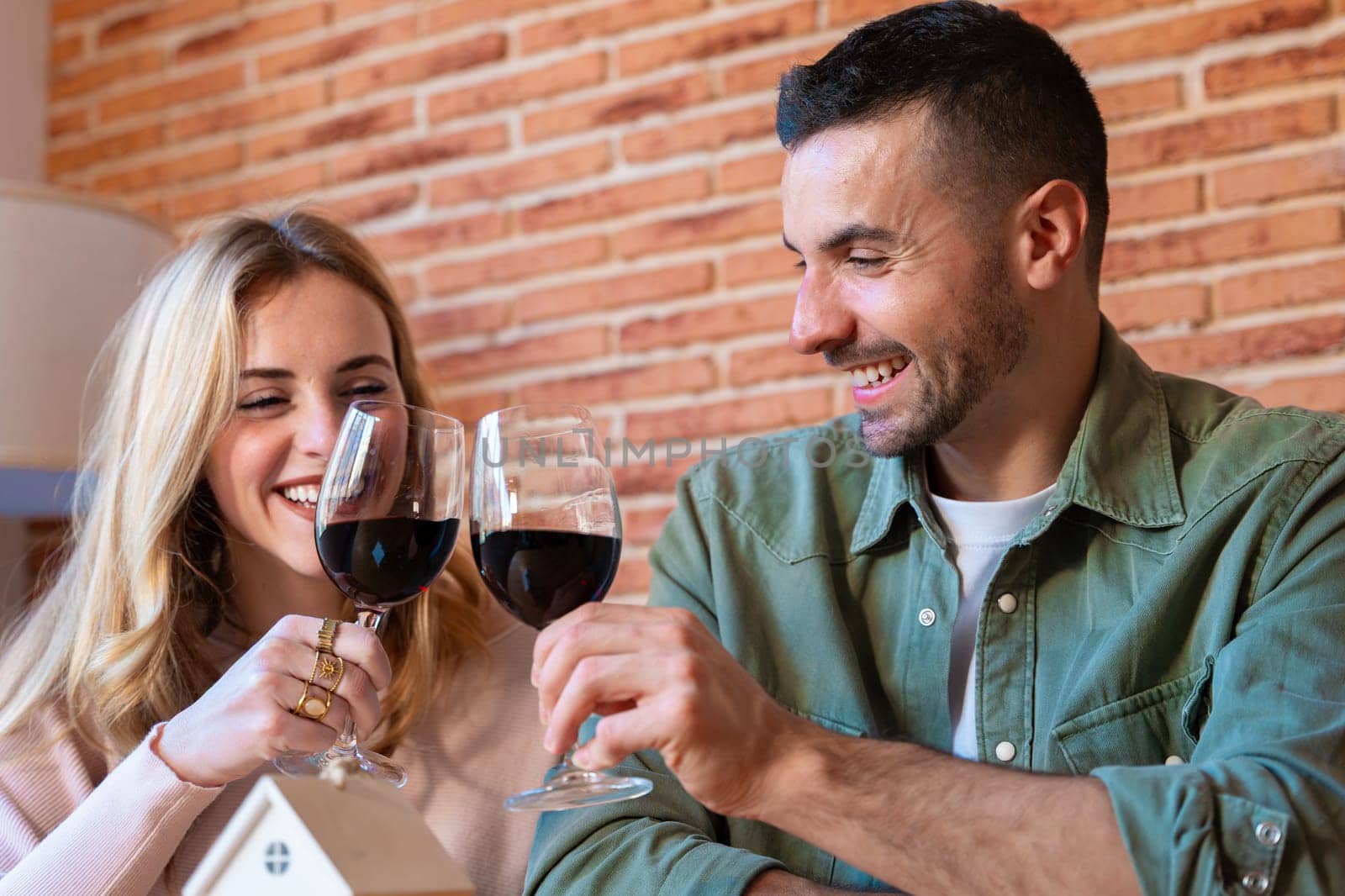 Smiling couple toasting with red wine while celebrating their new home. A miniature house and keys on the table symbolize the beginning of a new chapter in their life together.