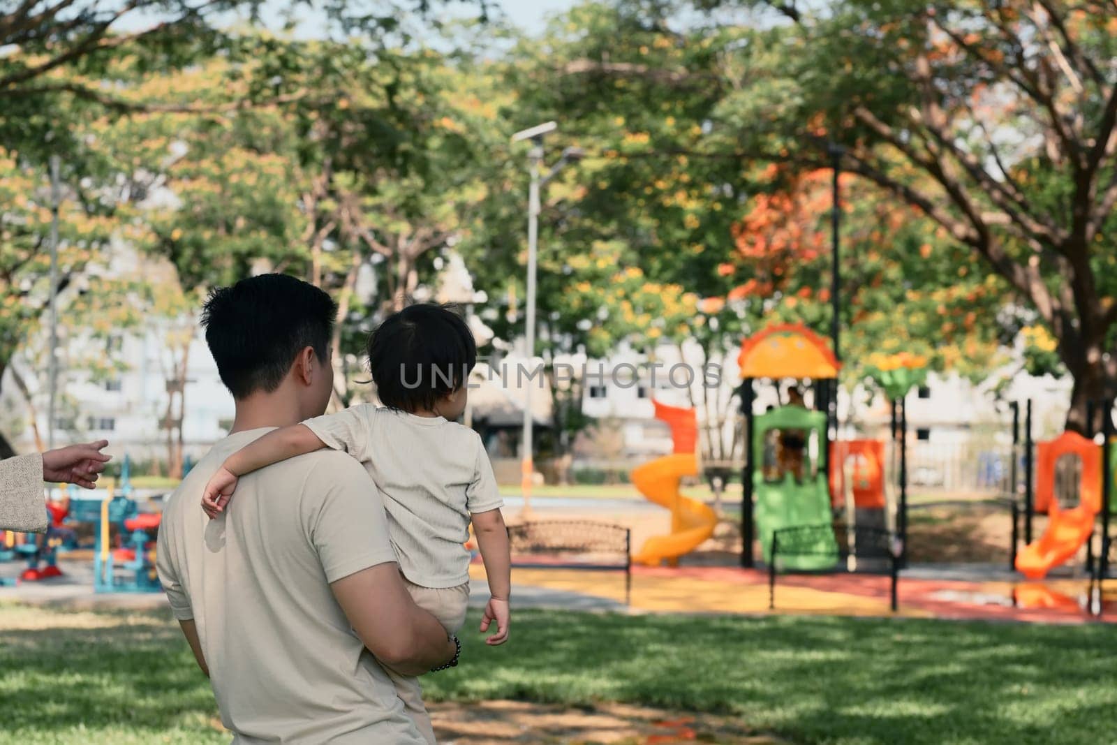 Father carries his toddler while walking toward a vibrant play area in a park.