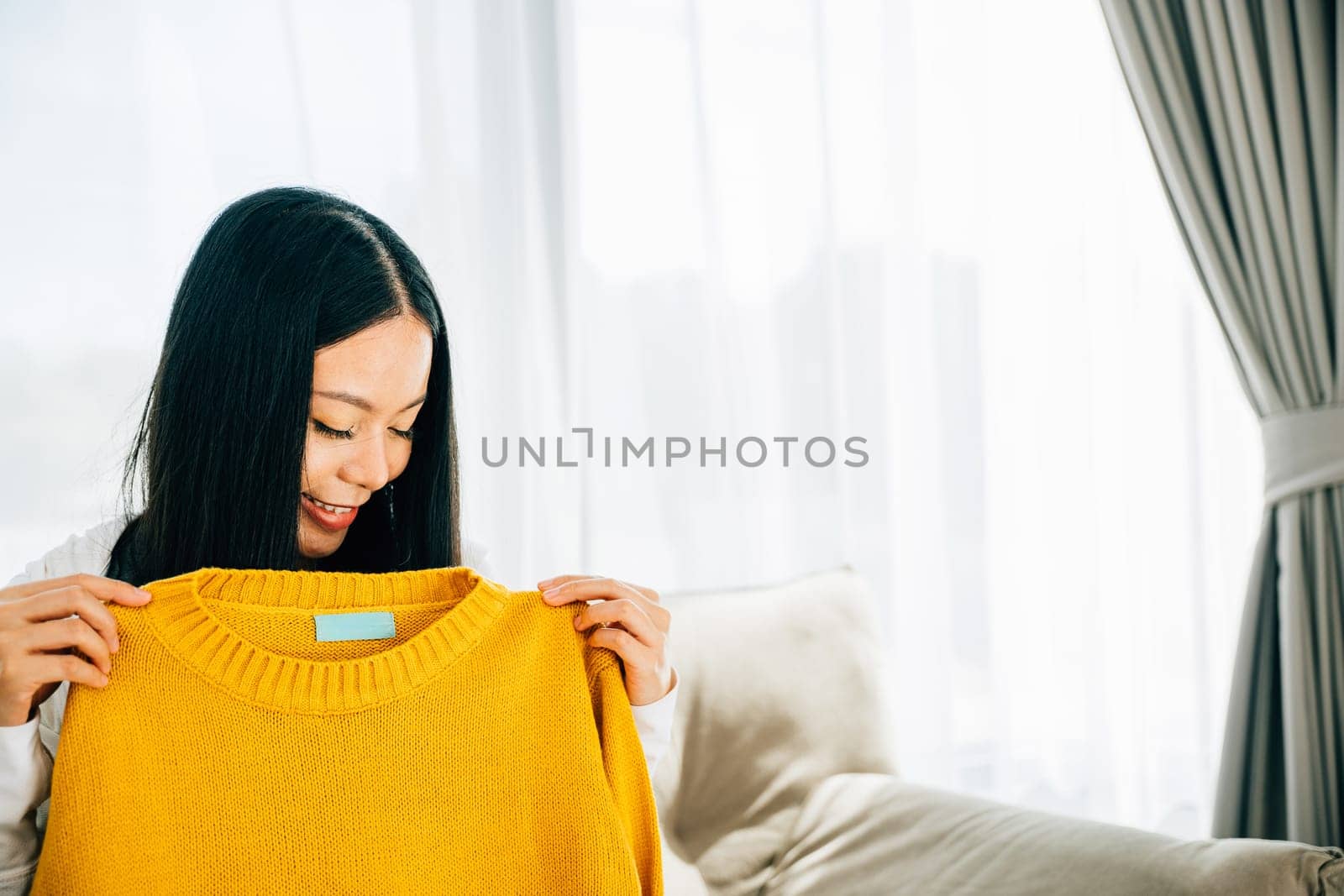 Excited Asian woman unpacks a shirt from a cardboard box smiling at home. Delighted customer unboxes online shopping package showcasing new clothes. Delivery and shopping concept.
