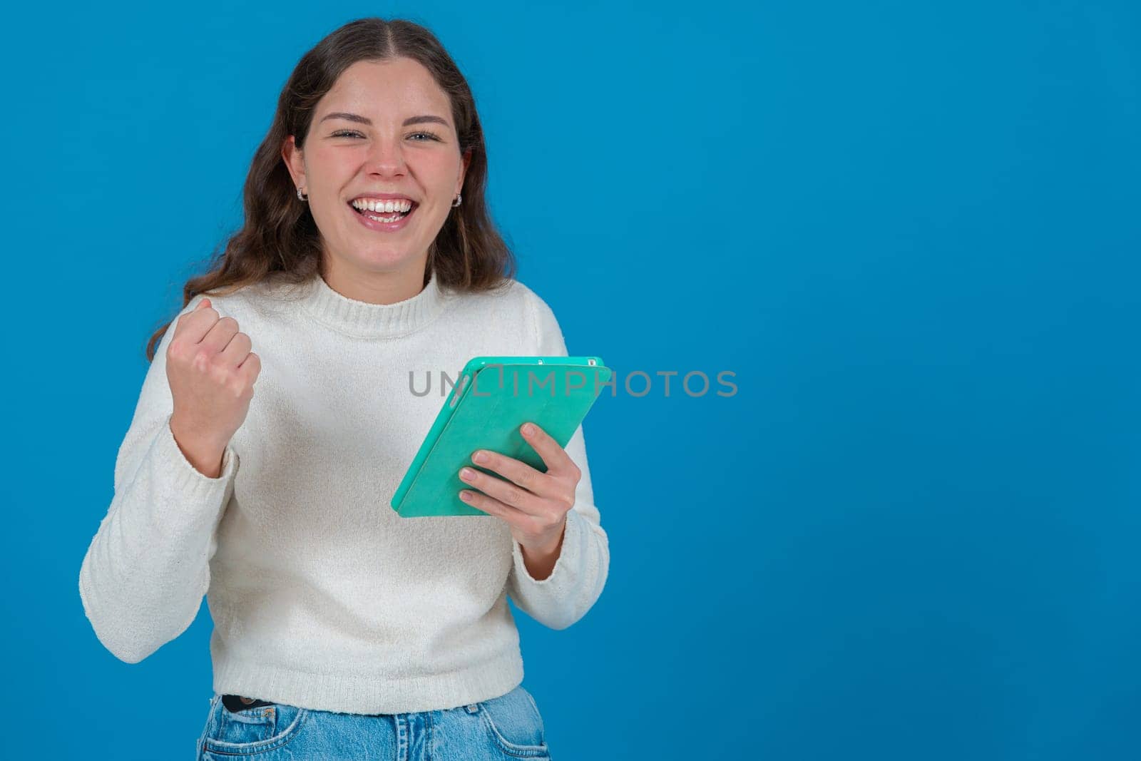 young woman in a white sweater raising her fist in celebration while holding a green tablet and smiling at the camera, against a blue background, symbolizing success, joy, and digital life. Copy space