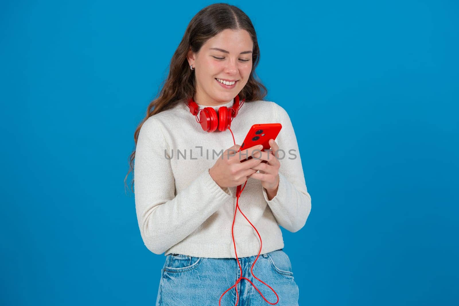 Happy young woman wearing red headphones around her neck while using her red smartphone. She smiles as she types a message, standing against a bright blue background in a casual moment.