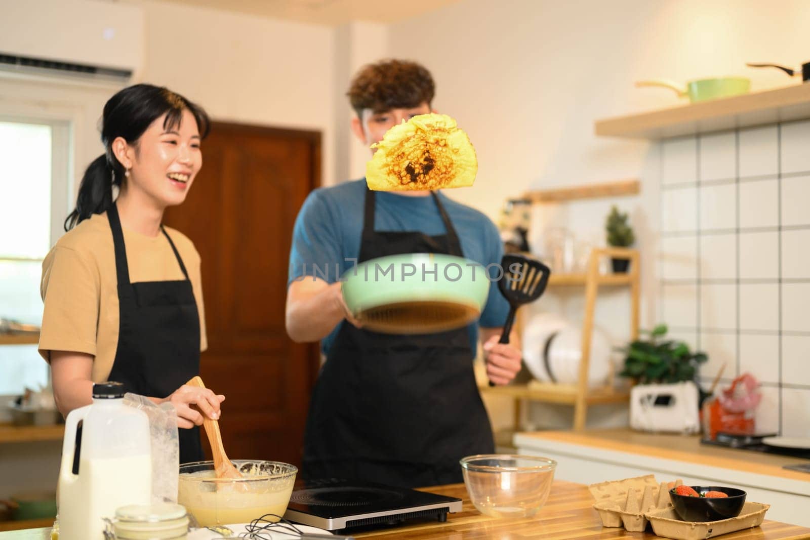 Playful couple flipping pancake, sharing laughter during home cooking.