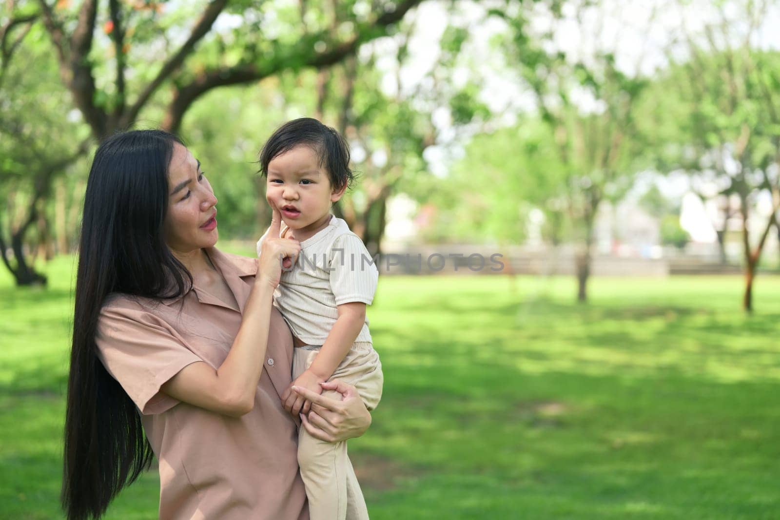 Asian mother and her toddler stroll through a lush green park.