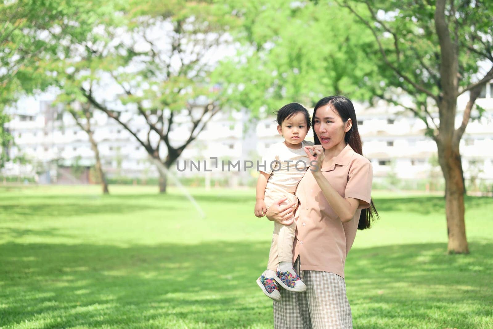 Asian mother and her toddler stroll through a lush green park.