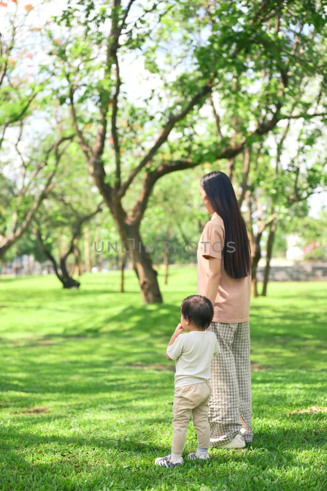 Asian mother and her toddler stroll through a lush green park.